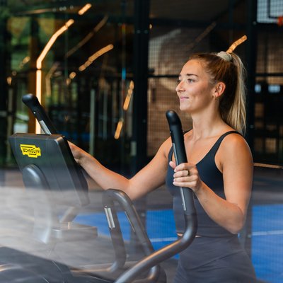 Eine Sportlerin nutzt den Crosstrainer im lichtdurchfluteten Fitnessbereich mit Blick nach draußen.