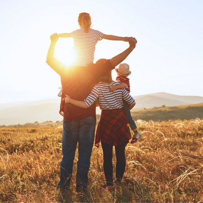 zwei Erwachsene, einer hat ein Kind auf den Schultern mit den Armen in der Luft, das andere Kind sitzt auf der Hüfte