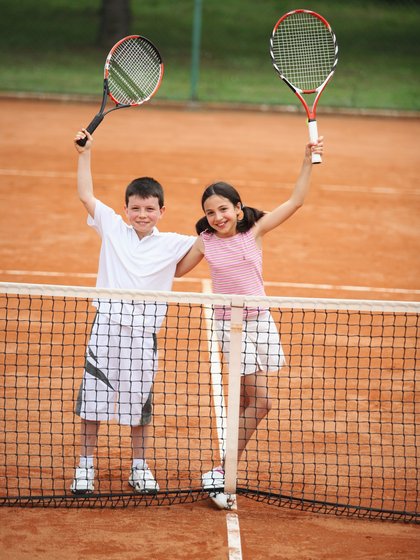 zwei Kinder auf einem Tennisplatz die Ihren Schläger in die Luft halten
