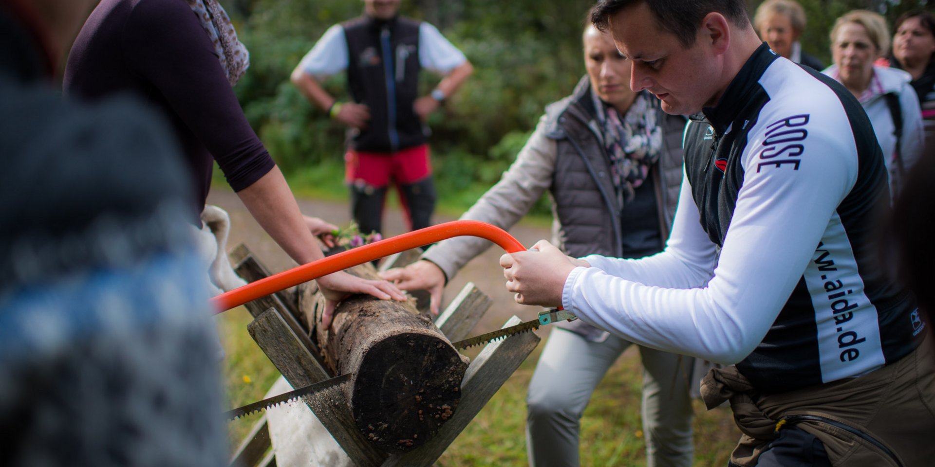 Mehrere Menschen stehen um einen Holzpfosten, der gerade von zwei Personen durchgesägt wird - Schwarzwaldgaudi