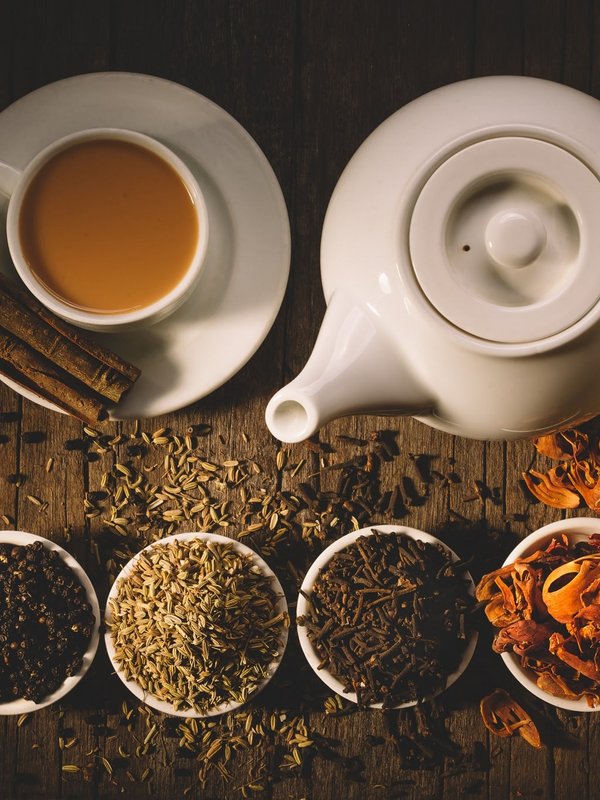 Eine weiße Teekanne mit Tasse und verschiedenen ayurvedischen Kräutern in Schalen auf dunklem Holz.