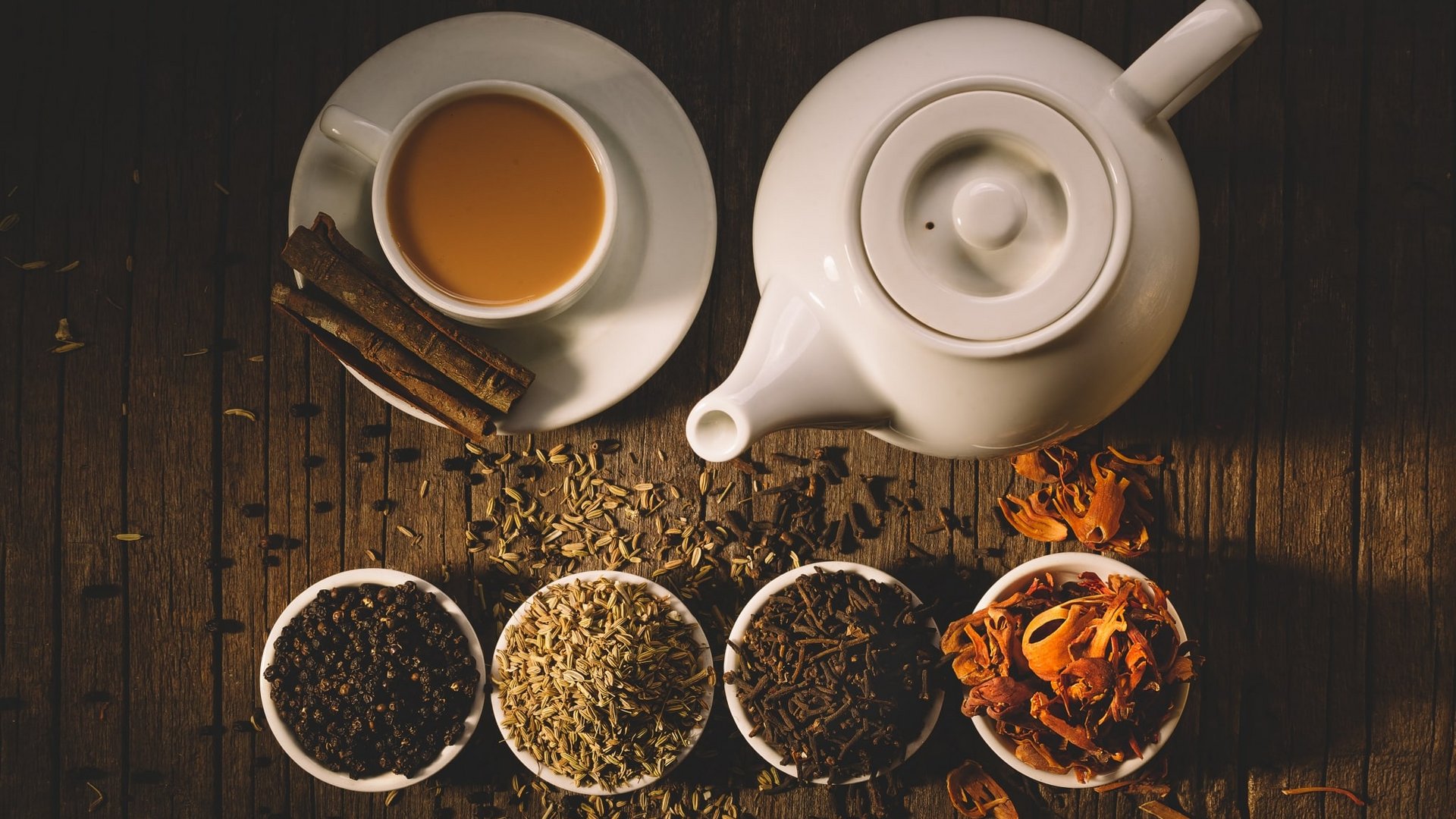 Eine weiße Teekanne mit Tasse und verschiedenen ayurvedischen Kräutern in Schalen auf dunklem Holz.