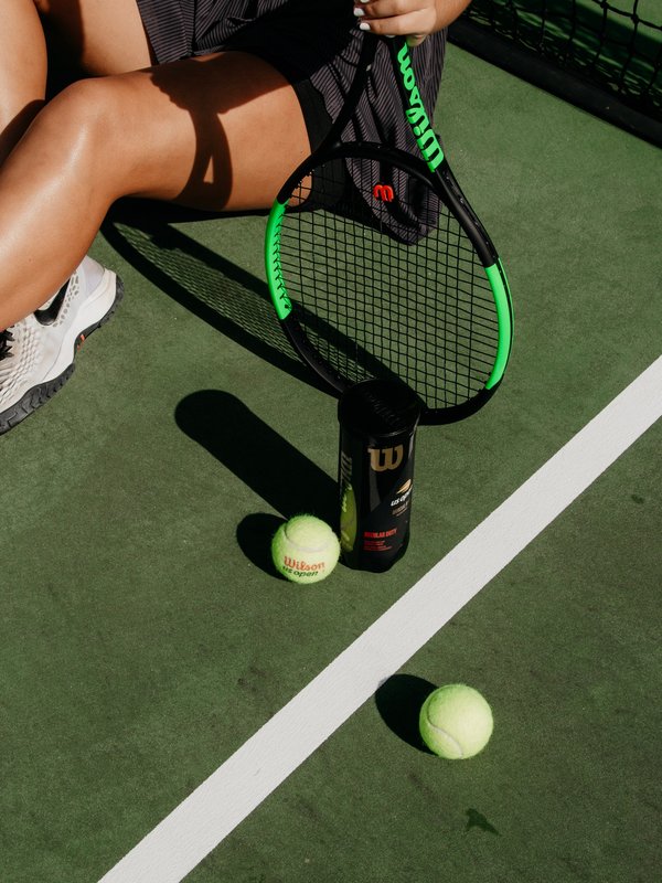 Beine einer Frau die auf einem Tennisplatz sitzt, neben Ihr liegen Tennisbälle und Schläger