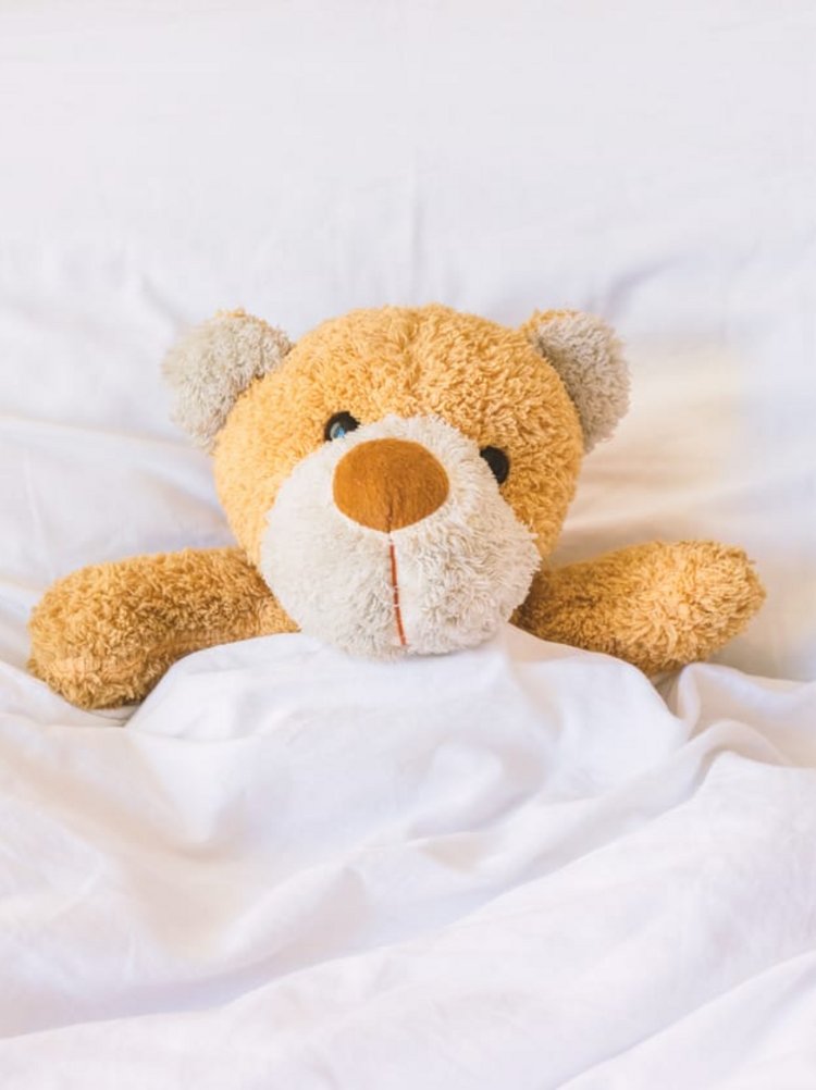 Ein flauschiger kleiner Teddybär liegt gemütlich unter einer weißen Bettdecke.