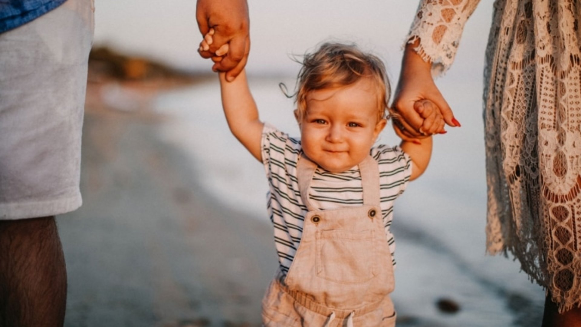 Ein kleines Kind macht seine ersten Schritte an den Händen der Eltern am sonnigen Seeufer.
