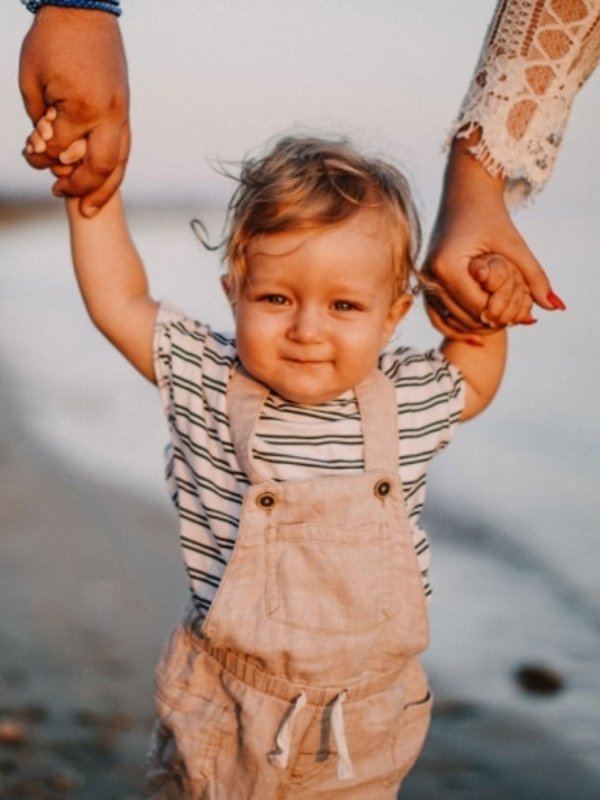 Ein kleines Kind macht seine ersten Schritte an den Händen der Eltern am sonnigen Seeufer.