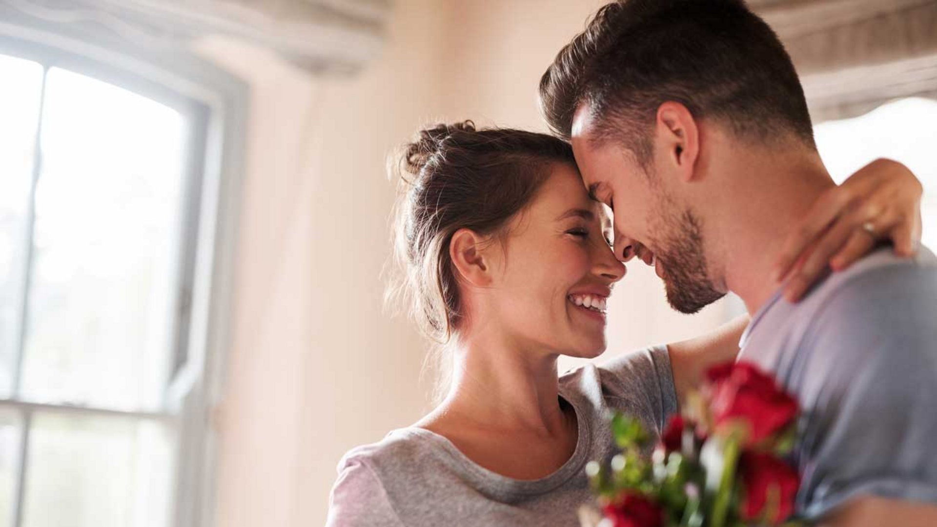 Ein Paar steht sich eng gegenüber; der Mann hält einen Strauß roter Rosen.