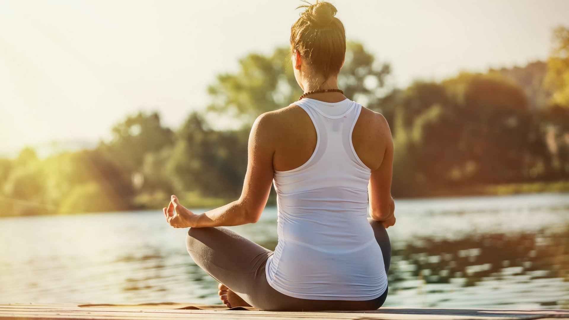 Eine Frau meditiert in der Yoga-Haltung am Ufer eines ruhigen Sees bei Sonnenuntergang.