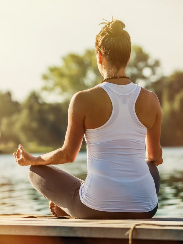Eine Frau meditiert in der Yoga-Haltung am Ufer eines ruhigen Sees bei Sonnenuntergang.