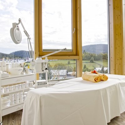 Ein lichtdurchfluteter Kosmetikraum mit weiß gedeckter Behandlungsliege und großem Panoramafenster im Hotel Vier Jahreszeiten.