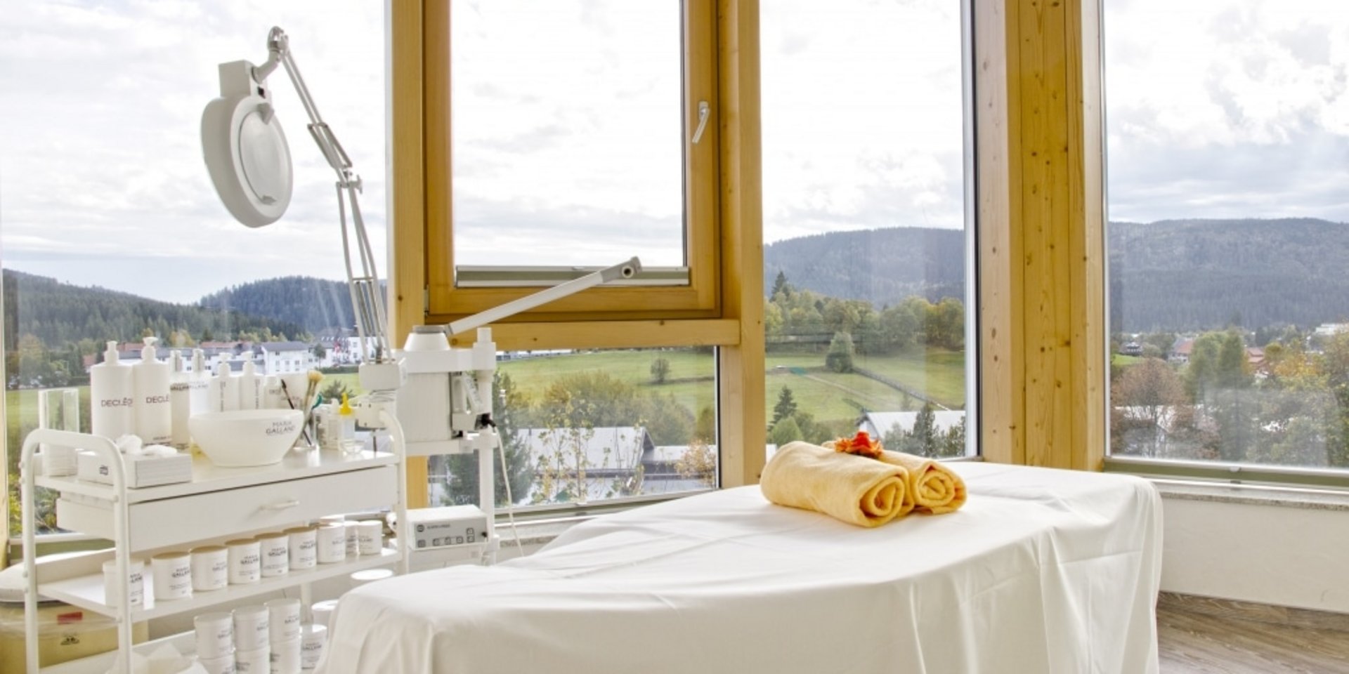 Ein lichtdurchfluteter Kosmetikraum mit weiß gedeckter Behandlungsliege und großem Panoramafenster im Hotel Vier Jahreszeiten.
