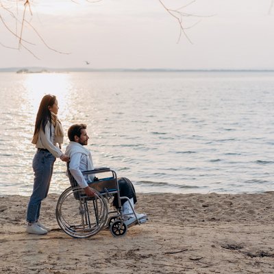 Eine Frau schiebt einen Mann im Rollstuhl am sandigen Ufer eines weiten Sees entlang.
